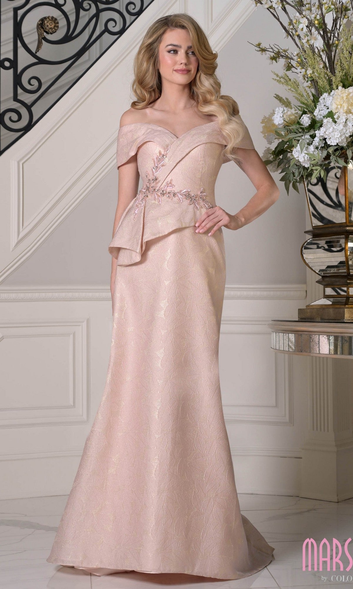 A woman stands indoors beside a table with flowers, wearing the blush Marsoni MV1397 long formal dress featuring an off-the-shoulder design, floor-length jacquard fabric, and floral embellishments at the waist.